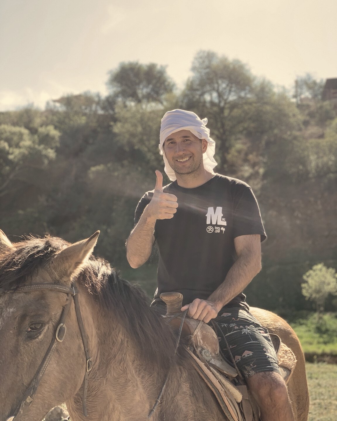 A smiling rider wearing a white head wrap gives a thumbs-up while seated on a brown horse.