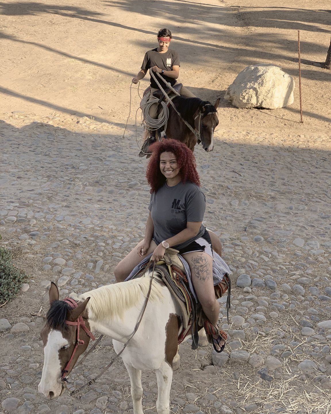 A smiling rider with curly red hair on a paint horse, with a rider coiling a rope behind her.